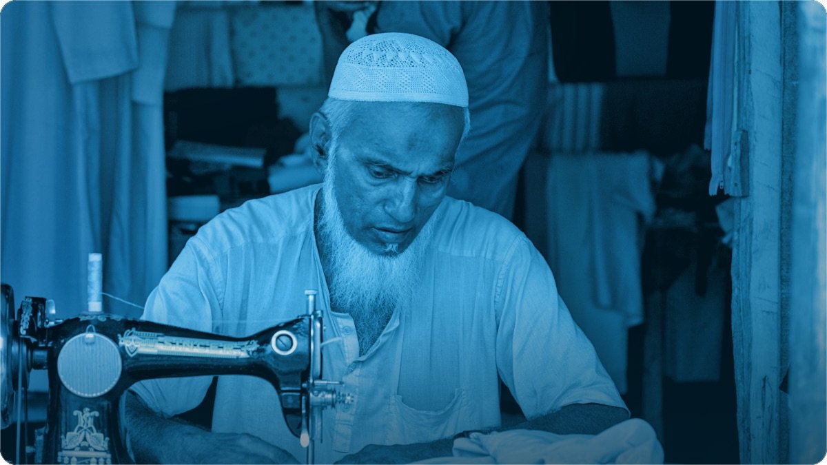 Chhoti dukan ke andar buzurg darzi sewing machine par kaam kar raha hai, peeche silay hue kapre latke hain.