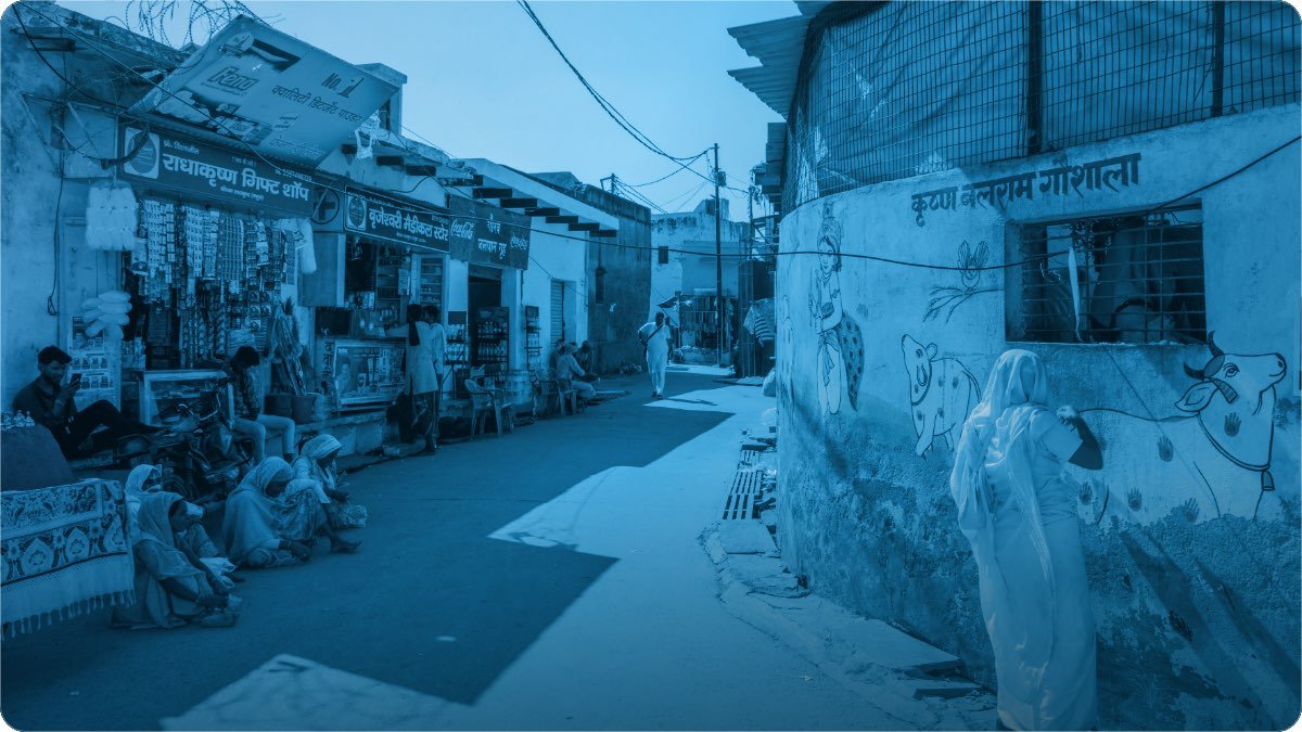 A small-town street lined with shops, signboards and goods spilling into the lane, people sitting outside.
