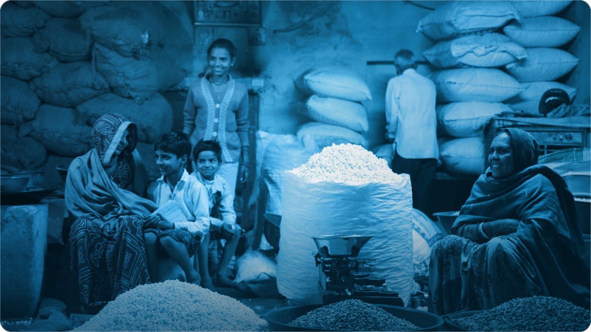 Vendors sit among open sacks of rice and pulses at a grain market, a weighing scale set between them.
