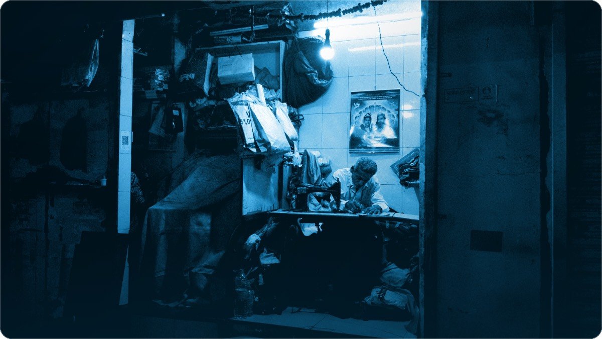 A tailor works at his sewing machine at night in a small brightly lit street-front workshop.