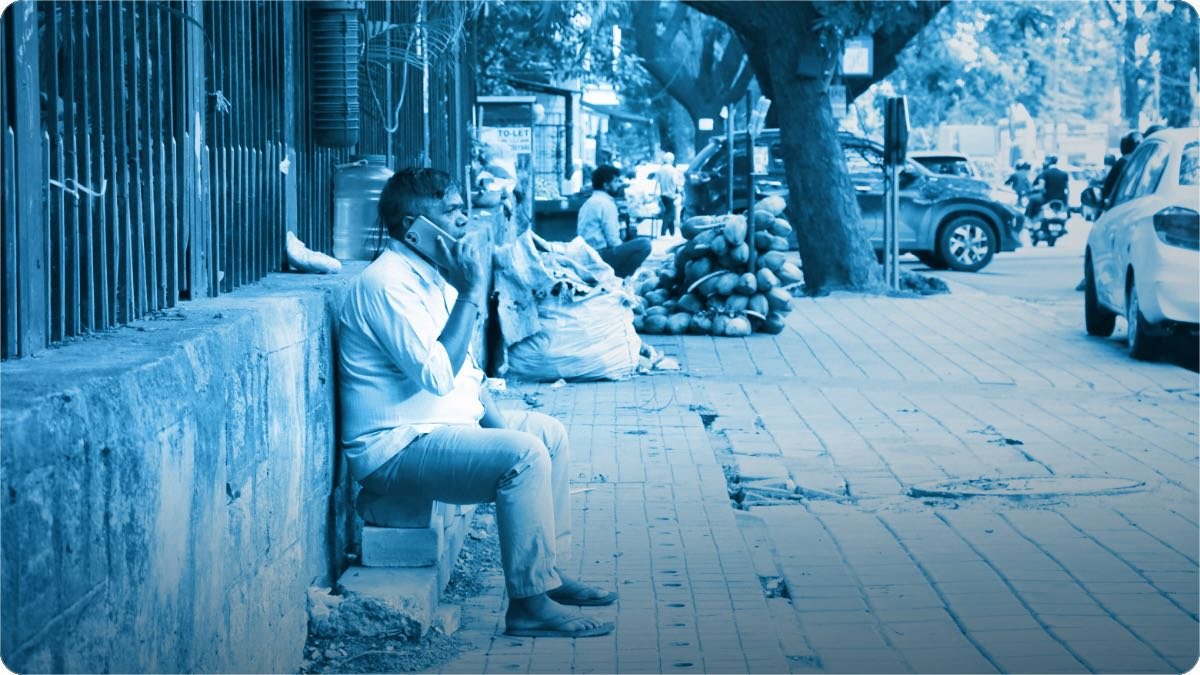A man sitting on a kerb on a busy street, talking on his phone beside a vendor’s sacks.