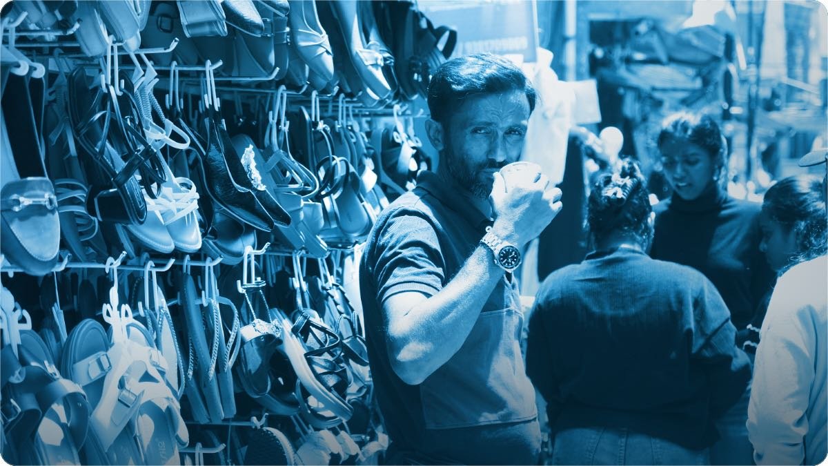 A footwear seller drinking tea beside a wall of hanging sandals while customers browse.