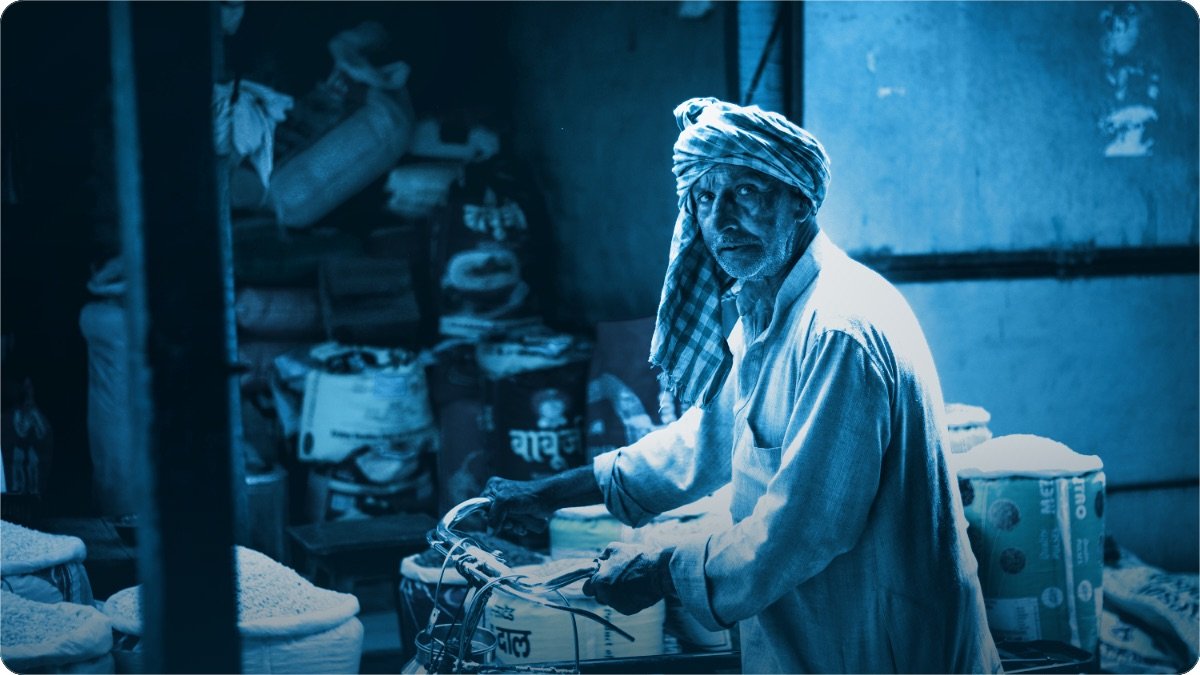 Pagri wala buzurg dukandar loaded cycle ke sath, bore aur carton se bhare huay store room mein khara hai.
