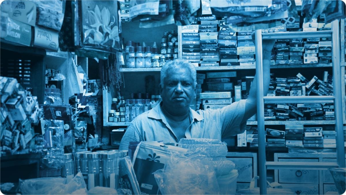 Ek dukandar apni bhari hui shelf ki taraf haath barha raha hai, chhoti dukan har cheez se bhari hai.