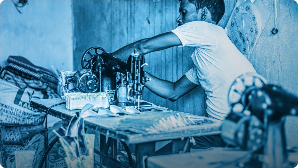 Darzi apni workshop mein pairon wali silai machine par kaam kar raha hai, mez par kate hue kapre rakhe hain.