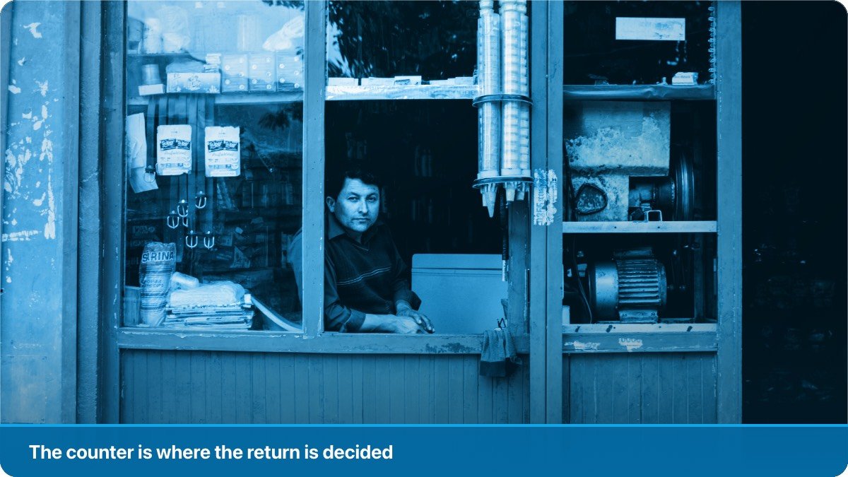 A shopkeeper at the open counter window of his small shop, goods stacked on the shelves behind him.