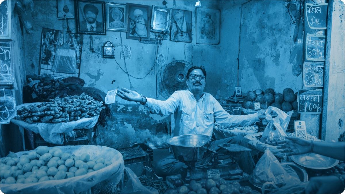 A shopkeeper talking with a customer across a counter piled with vegetables.