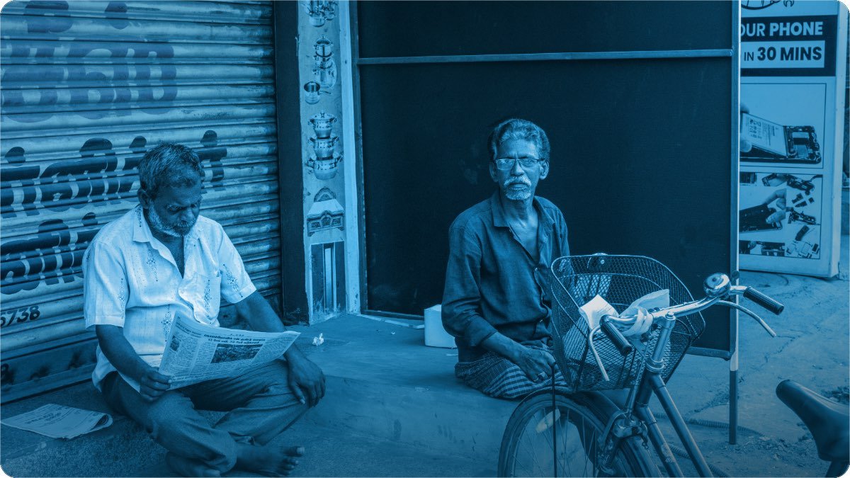 Band shutter wali dukan ke bahar do buzurg baithe hain, ek akhbar parh raha hai, paas cycle khari hai.