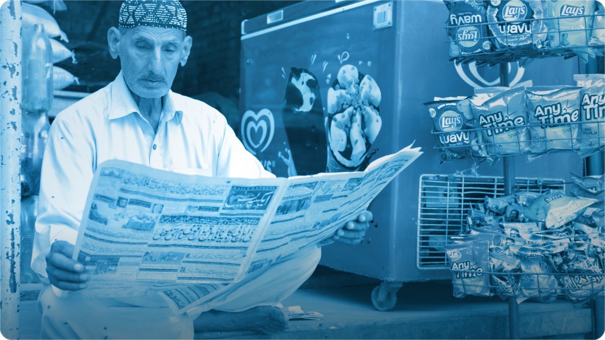 Umar rasida dukandar khamosh dukan mein Urdu akhbar parh raha hai, paas freezer aur latke hue chips ke packet.