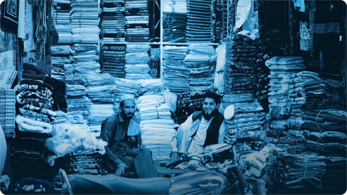 Two shopkeepers sitting in a small cloth shop stacked floor to ceiling with folded fabric.