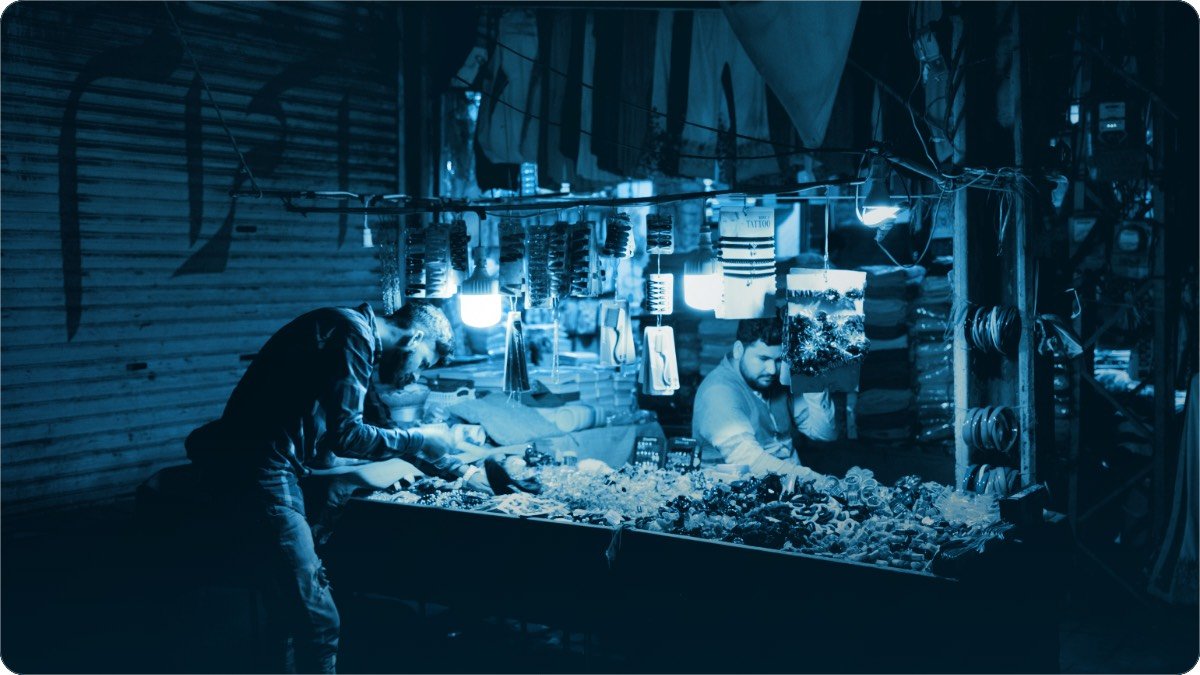 Do aadmi raat ke bazaar mein aik hi thele par kaam karte hue, bulb ke neeche maal phaila hua.