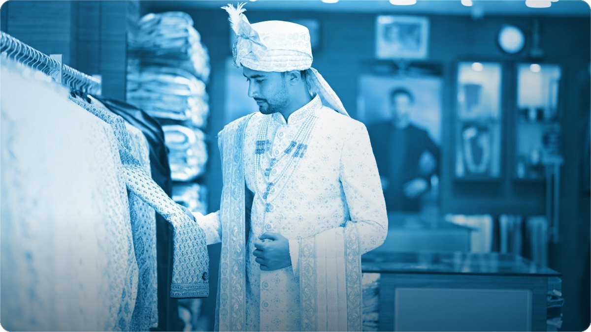 Kadhai wali sherwani pehne aadmi kapron ki dukan mein rack par tange suit dekh raha hai.