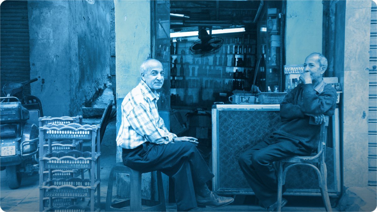 Two men, one older and one younger, sitting at the front of a small shop stacked with crates and shelves.