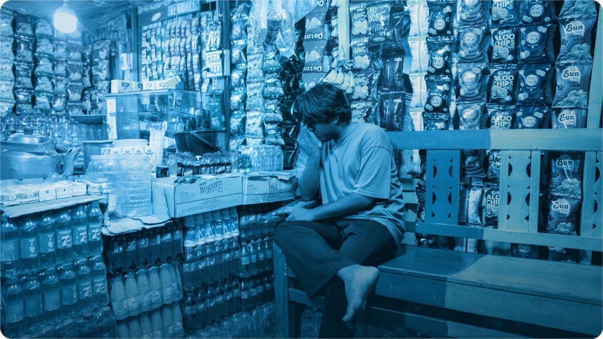 A young shop helper sits on a bench inside a small snack shop, its walls stacked with crisp packets and bottles.