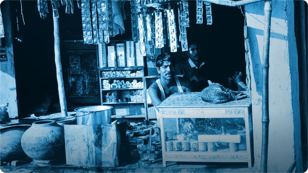 Gaon ki dukan, latki hui sachet ki lariyan aur sheeshe ka showcase, counter ke peeche do bande baithe hain.