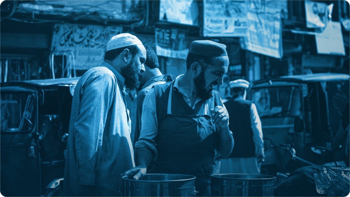 Two men standing close together at a street stall, talking while one holds a small container.