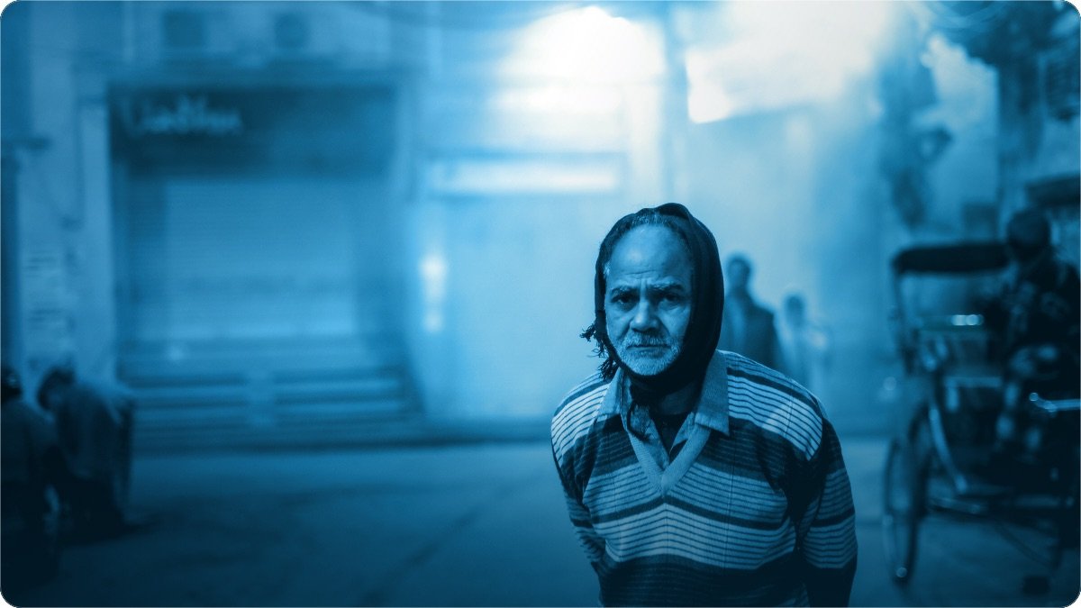 An older man walking a misty street at dusk past a shuttered shop, a cycle rickshaw waiting behind him.