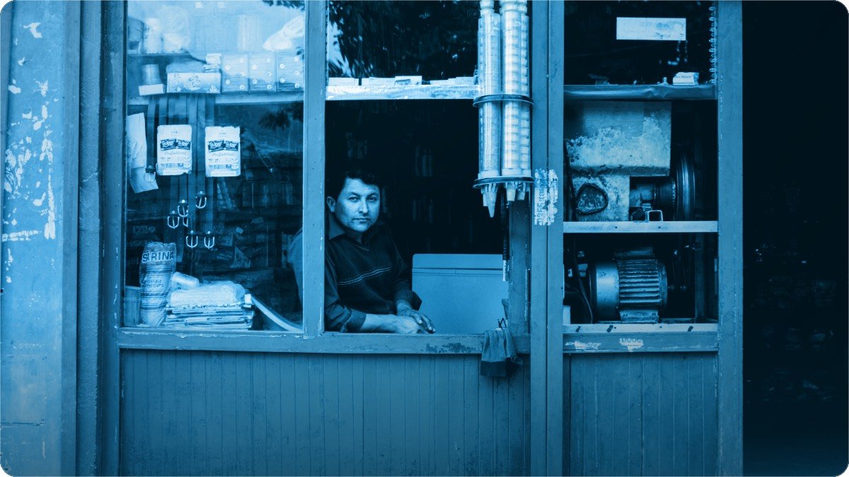 A shopkeeper sitting at the open window of his shop, goods stacked on the shelves behind him.