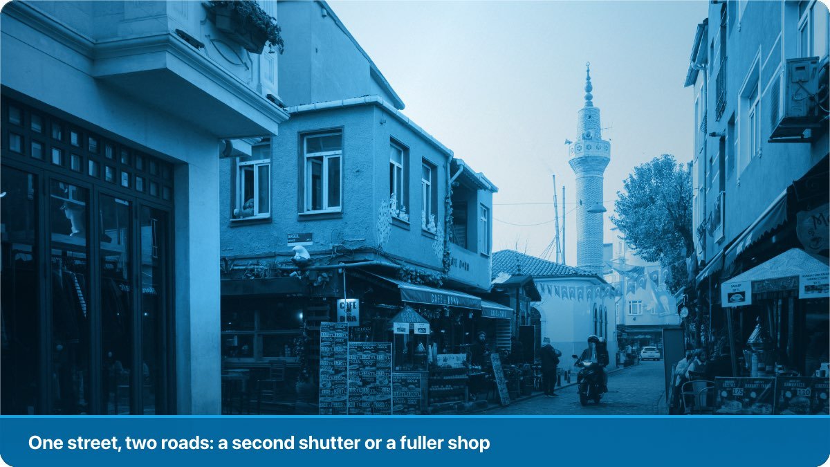 A narrow street lined with small shops on both sides, awnings out and a motorcycle passing.