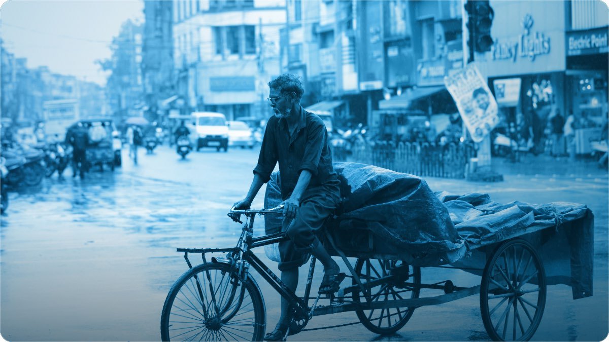 A man pushes a handcart of goods through a wet bazaar street, the load tied down under a tarpaulin.