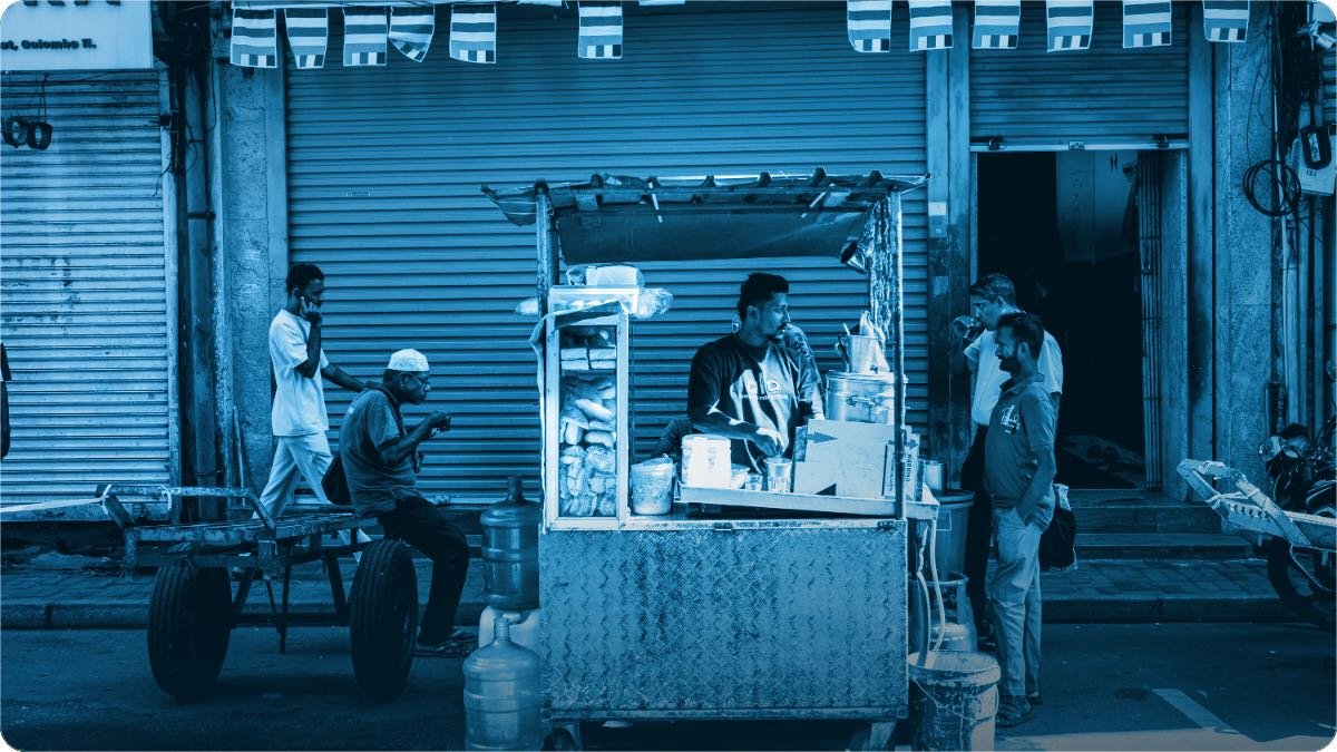 A street food cart lit up at dusk with customers standing around it and closed shutters behind.