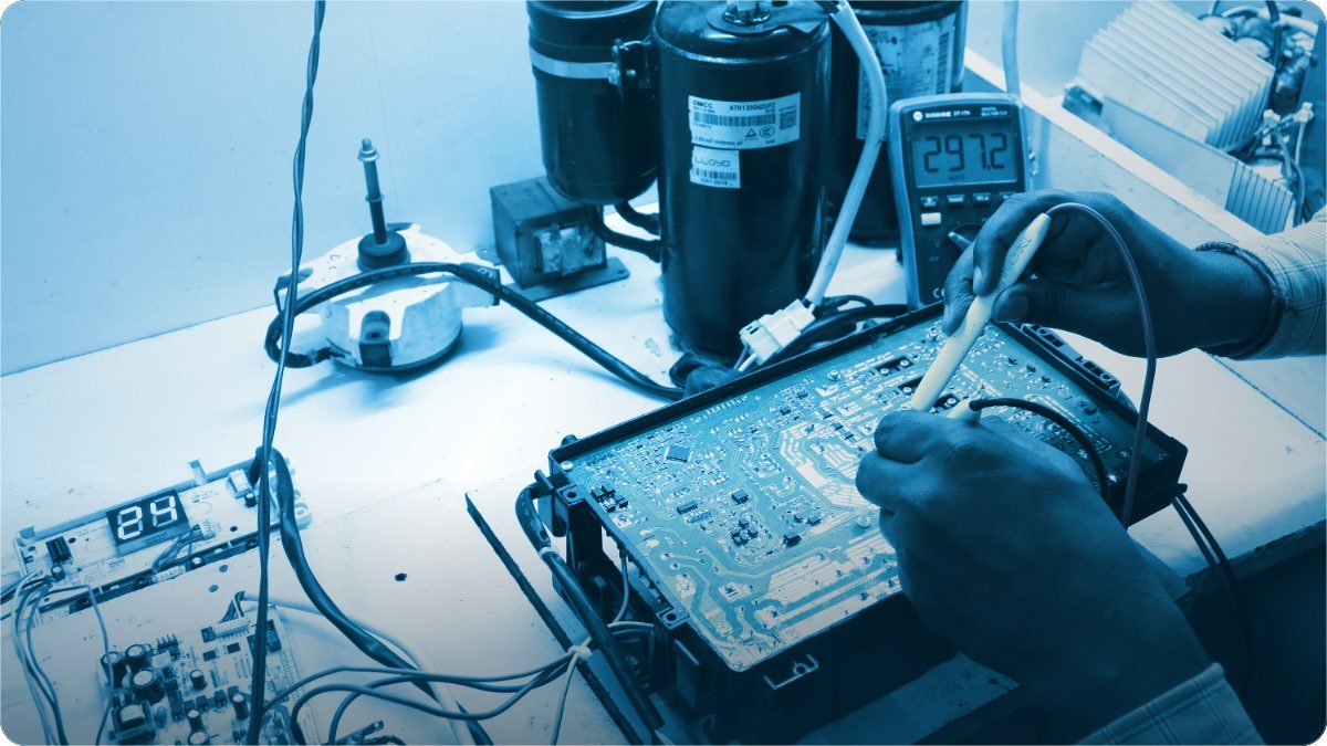 Hands test an appliance circuit board with a meter, compressors and spare parts laid out on the workbench.