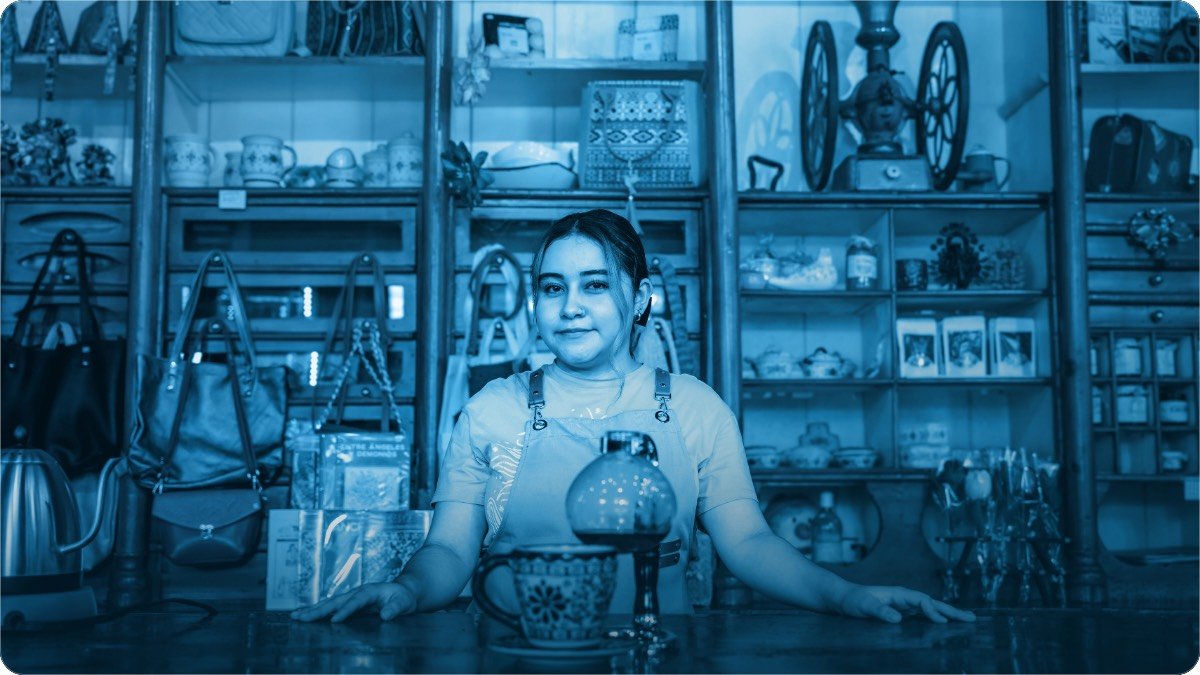 A shopkeeper standing at a busy counter with stocked shelves behind.