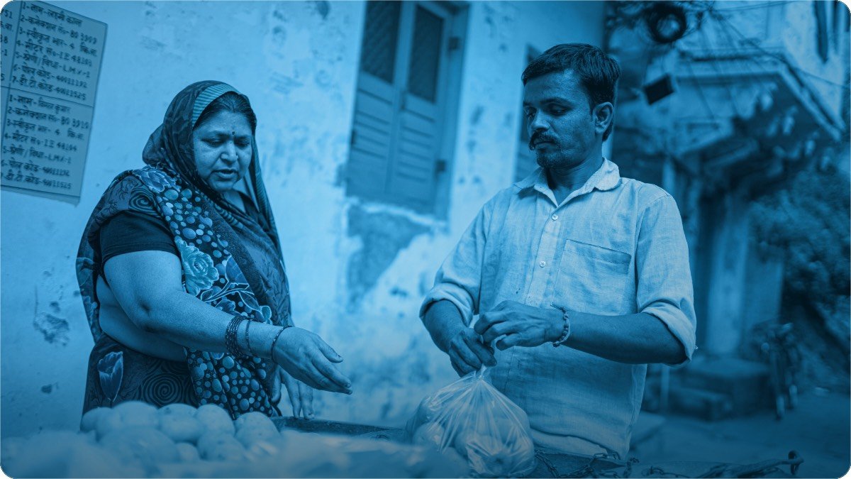 Sabzi ke thele par dukandar aur customer sauda tay kar rahe hain, darmiyan mein bhara hua shopper.