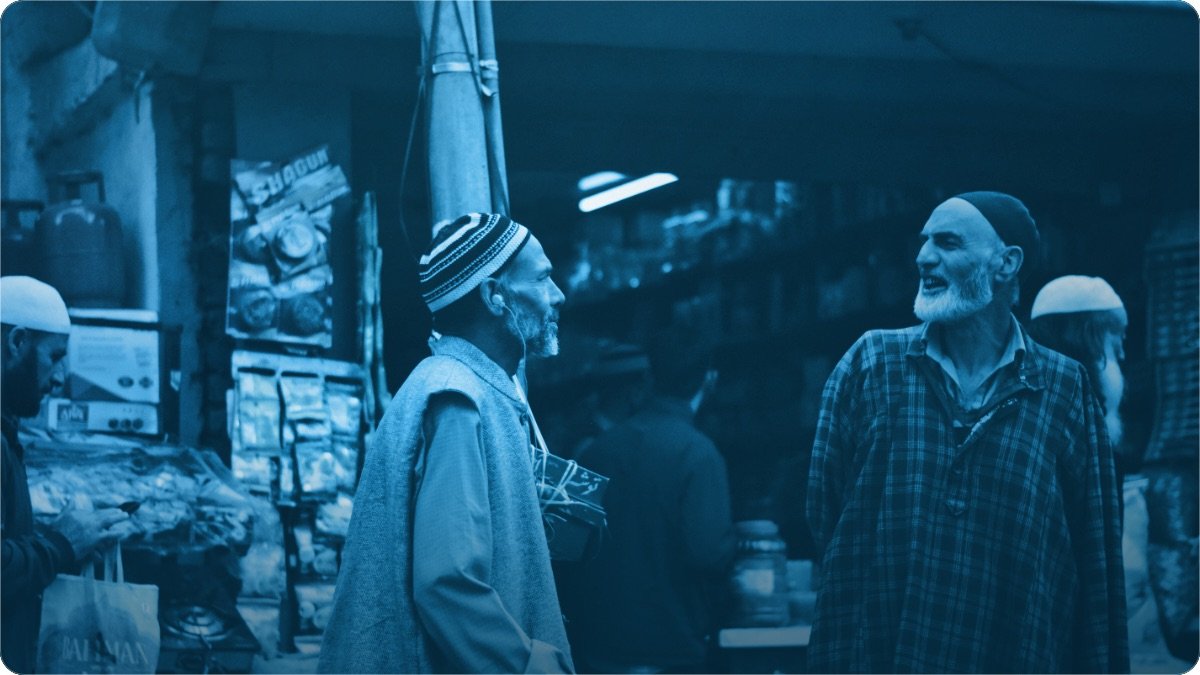 Two shopkeepers standing face to face in a busy market lane, talking over a deal.