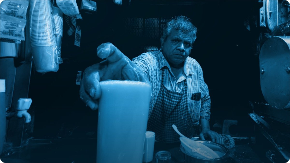 A stall owner handing a filled glass across the counter of his small roadside shop.