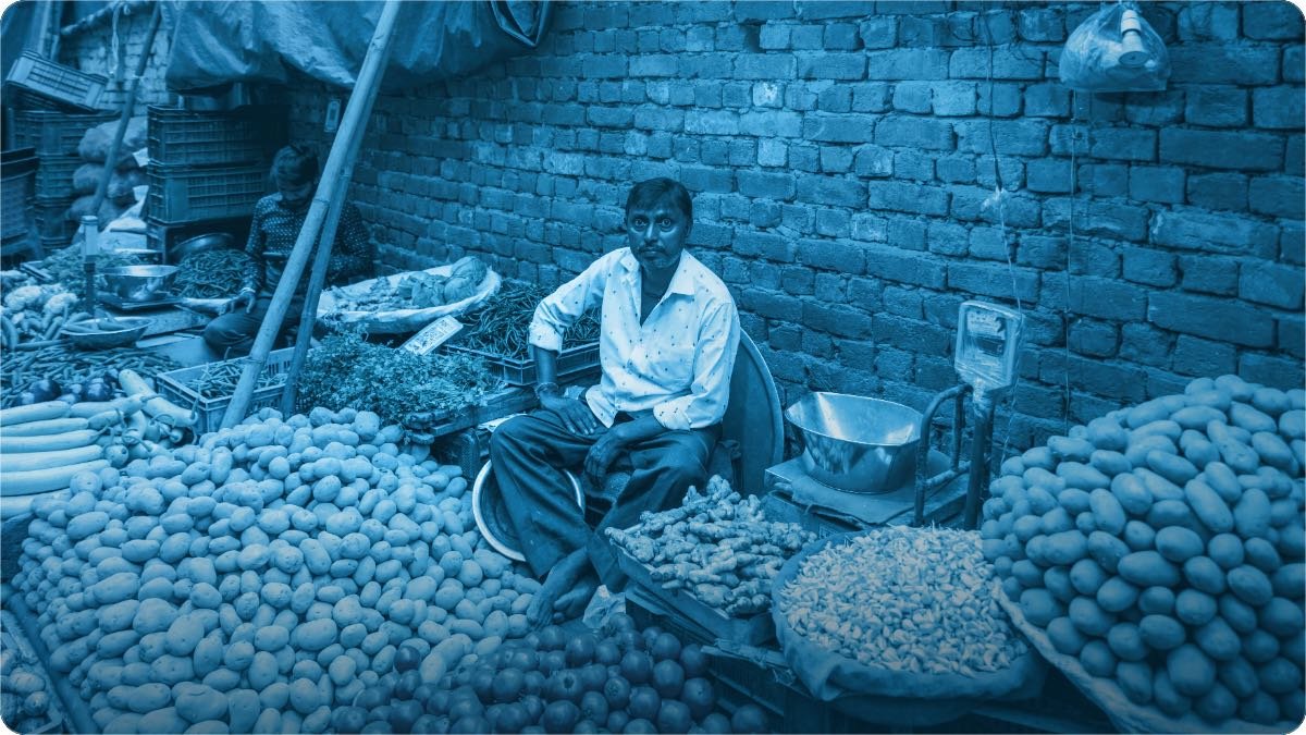 A vegetable seller sitting among his piles of potatoes and greens at a market stall.