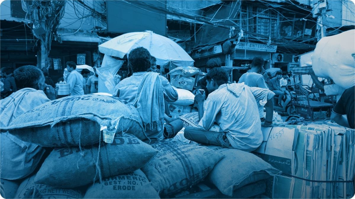 A crowded South Asian wholesale market, sacks of goods stacked in front and traders working behind them.