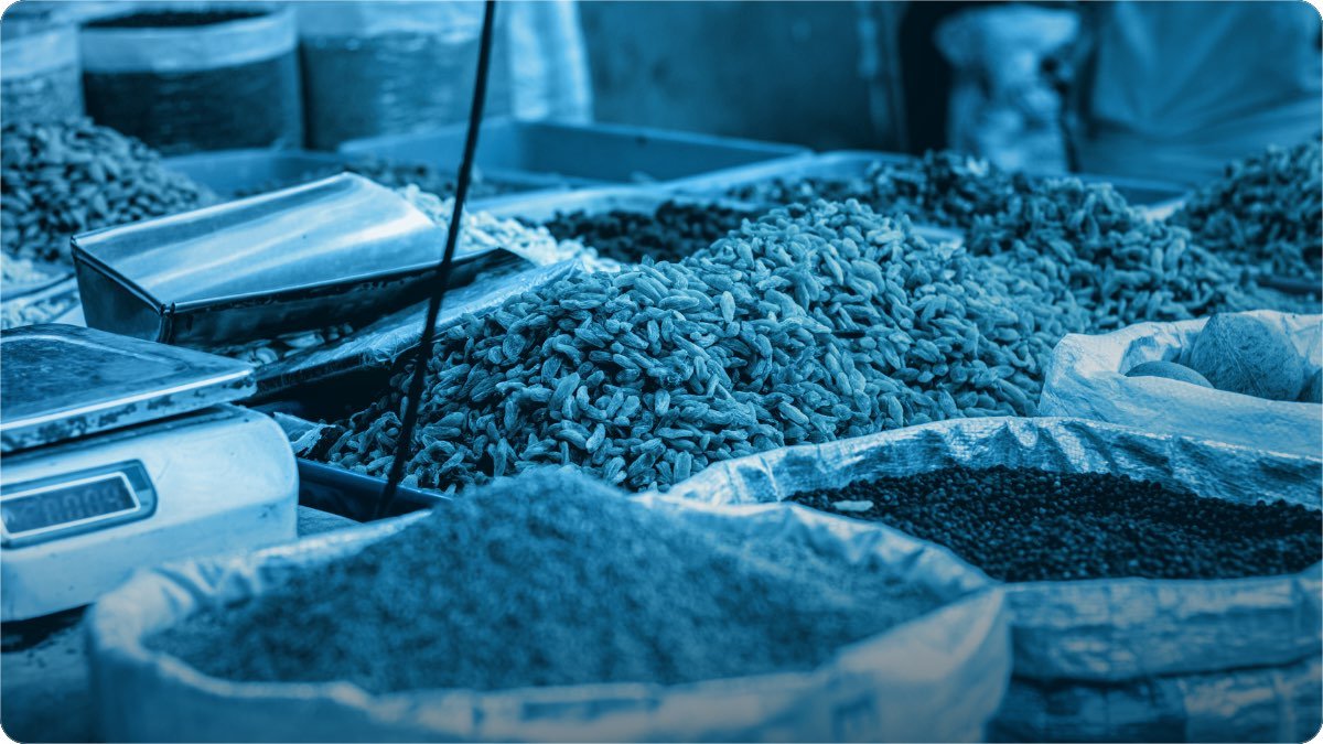 Open sacks of grain and spices at a wholesale stall, with a weighing scale and metal scoops beside them.