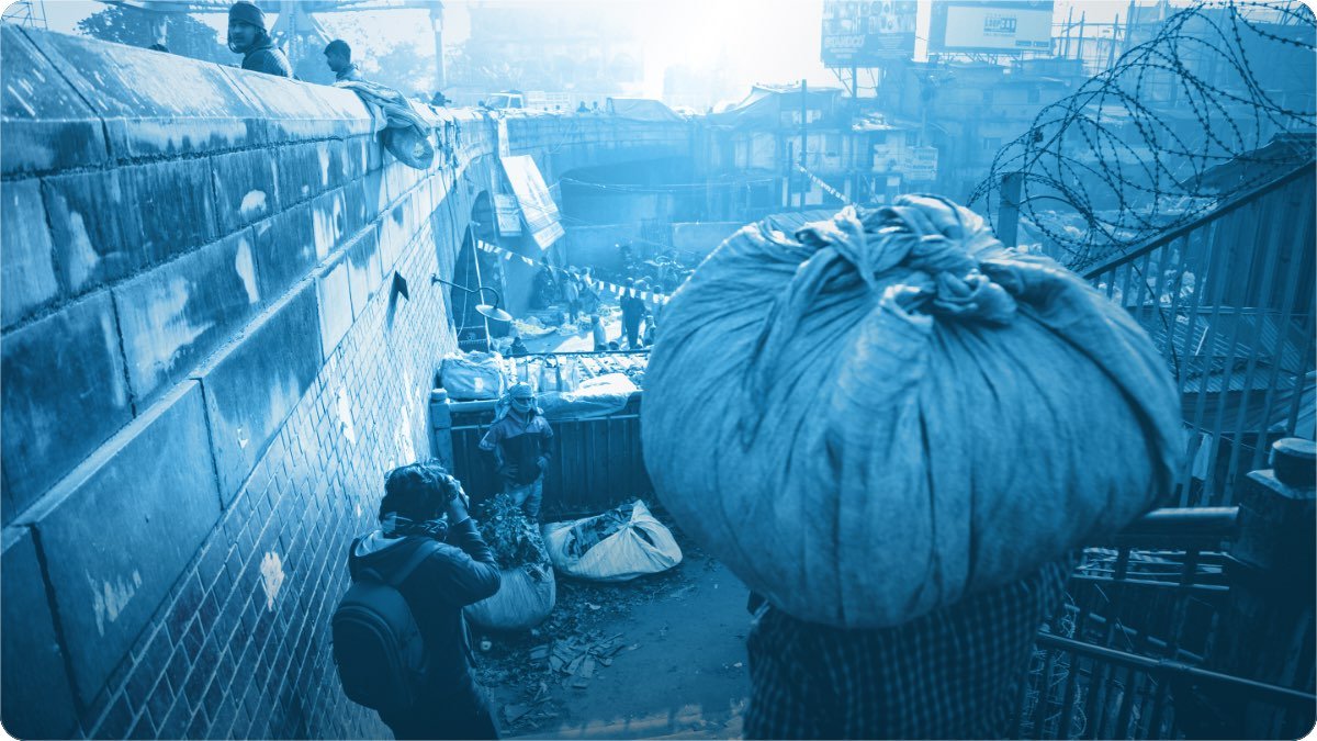 A porter carries a huge cloth bundle on his head through a crowded market lane packed with sacks.