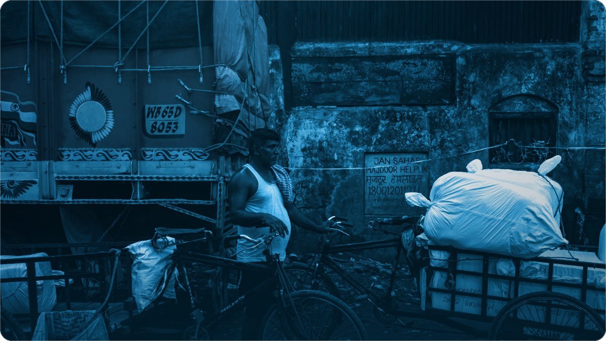 Goods being moved on a market street, a loaded truck behind and sacks stacked high on a hand cart.