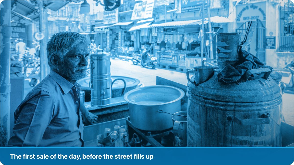 A tea stall owner waiting at his counter in the morning, kettles and pots ready, the street still empty.