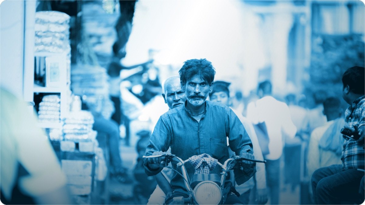 A man rides a motorcycle with a second rider through a crowded South Asian market street.