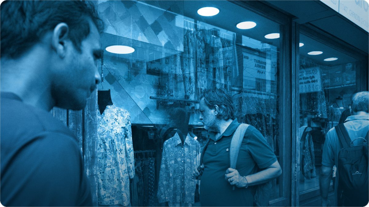 Masroof bazaar mein kapre ki dukan ke shishe ke saamne khare log, andar jore lagay hue.