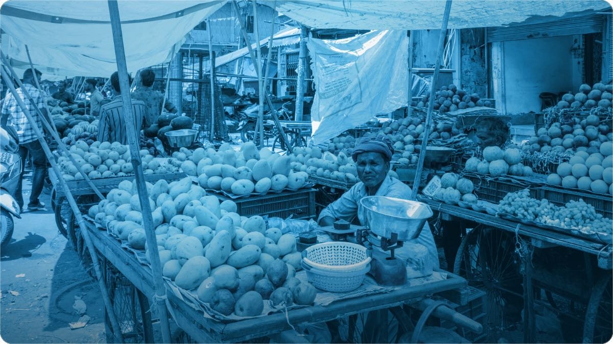 A fruit seller sitting at his roadside stall in South Asia, produce piled on the tables around him.