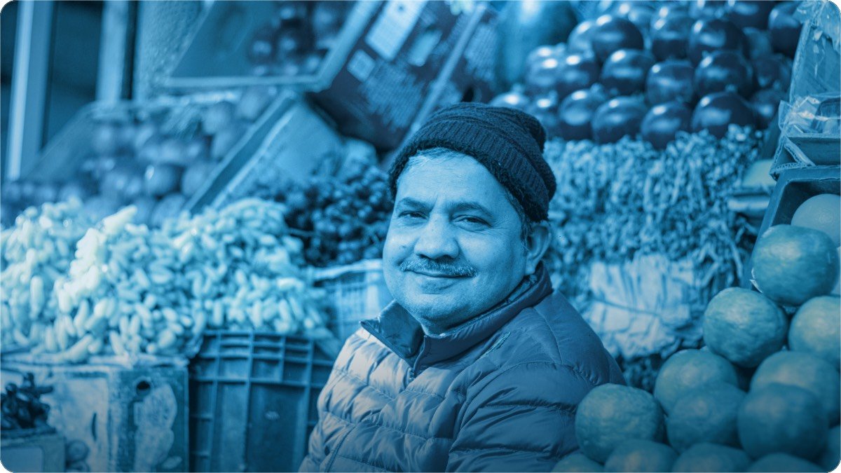 A shopkeeper in a woollen cap sits at his fruit stall, bananas and oranges stacked around him.