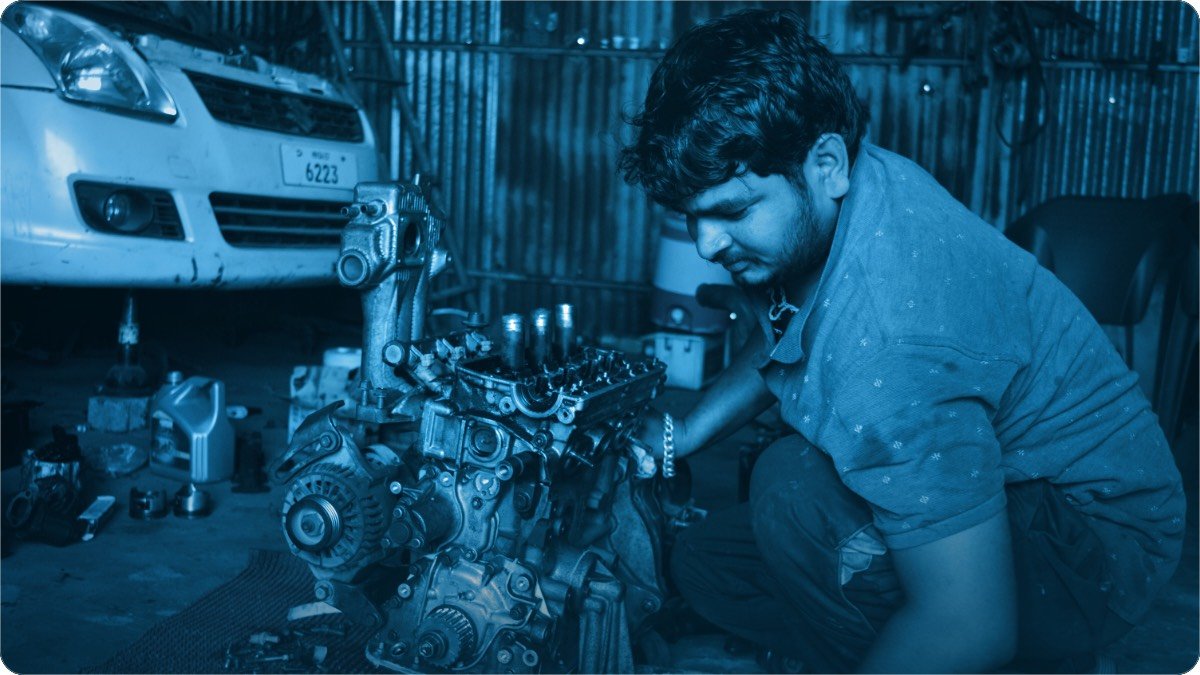 Nojawan mechanic choti workshop mein khuli hui engine par jhuka hua, peechhe gaari khari hai.