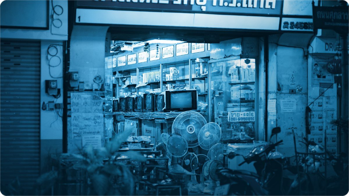 A lit street electronics shop at night with fans, a television and boxed goods stacked at the front.
