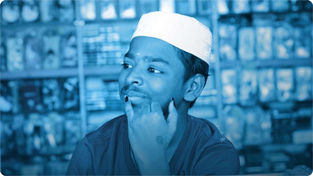 A young shopkeeper resting his chin on his hand, thinking, with stocked shelves behind him.