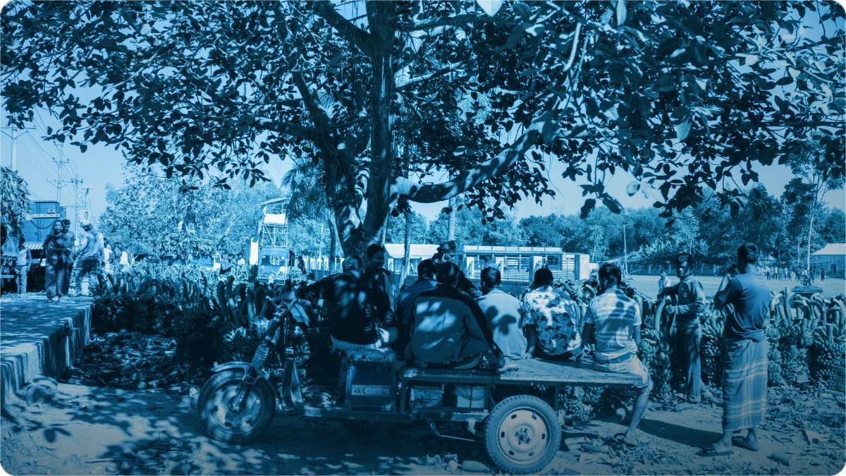 Traders sitting together on a cart at a village market, stacks of bananas piled around them.