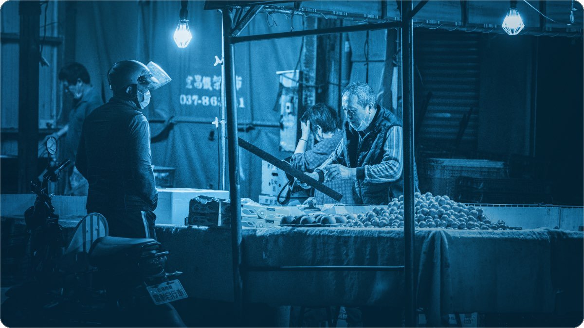 A vendor serving a customer at a lit fruit stall in the evening market.