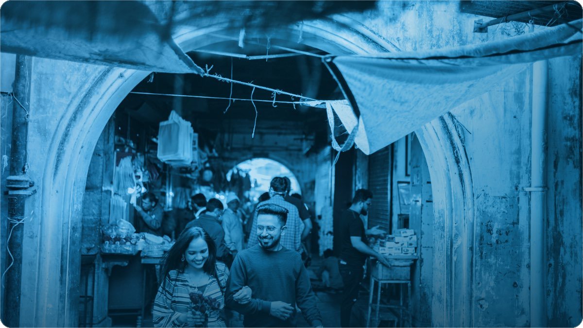 Bazaar ki dhaki hui gali mein chalta hua jora, peechhe dono taraf market ke thele.