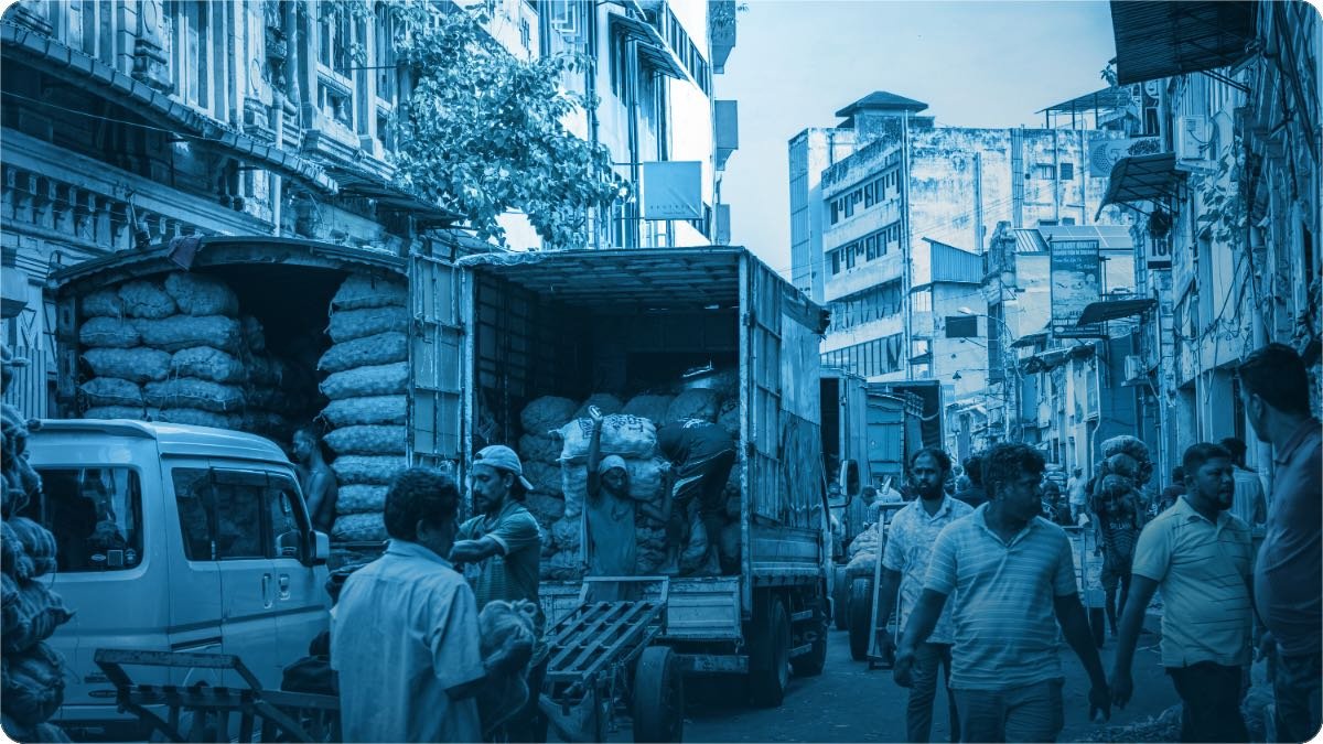 Mazdoor bheer wale bazaar mein truck se boriyan utaar rahe hain, dono taraf dukanein hain.