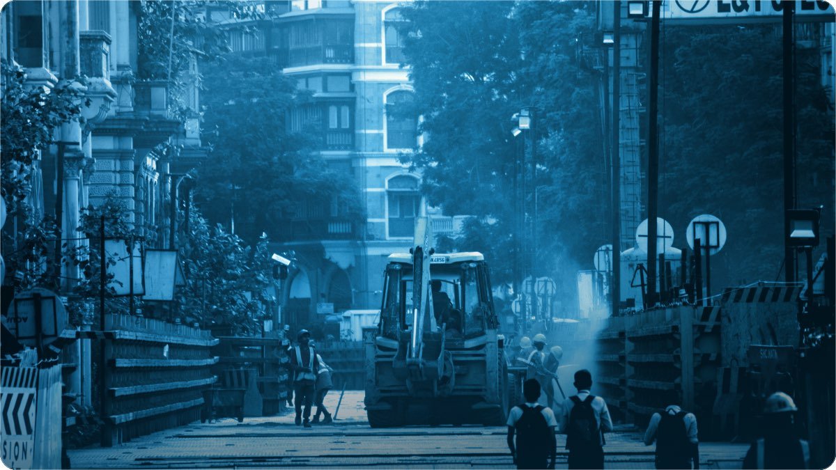 A city street dug open for construction, a digger and workers between the shops and old buildings.