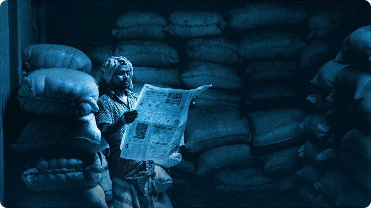 Ek aadmi kam roshni wale store mein anaj ki chini hui borion ke darmiyan akhbar parh raha hai.