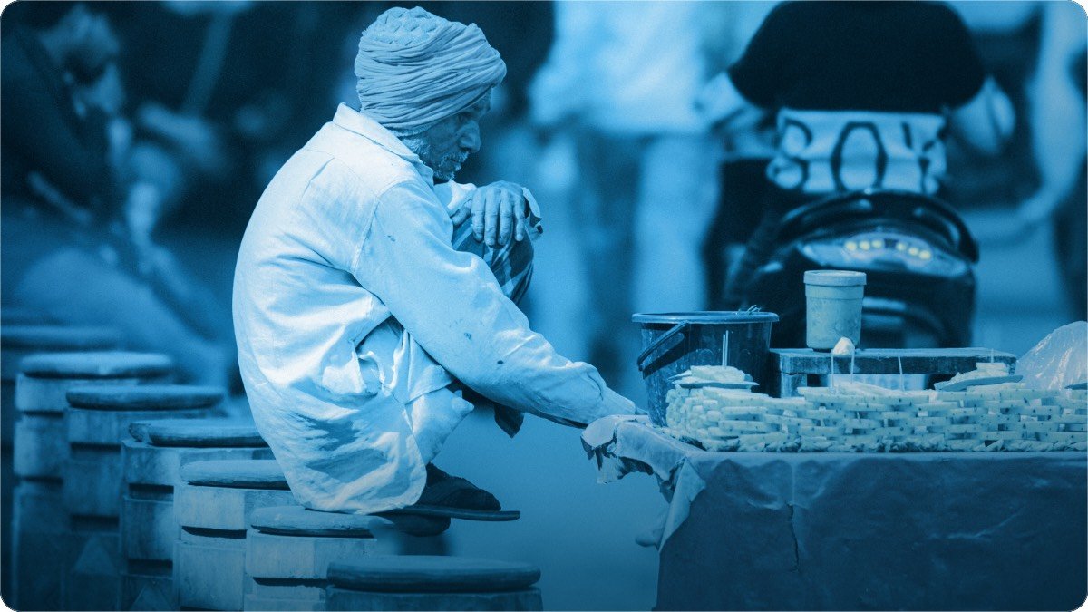 An elderly street vendor in a turban sitting quietly beside his small roadside stall.